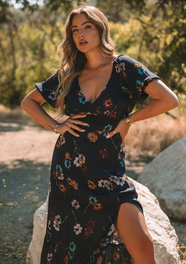 [Color: Navy/Mustard] A close up front facing image of a blonde model standing outside wearing a bohemian fall maxi dress in a navy blue and mustard yellow floral print. With short puff sleeves, a v neckline, a long flowy tiered skirt, and a side slit.