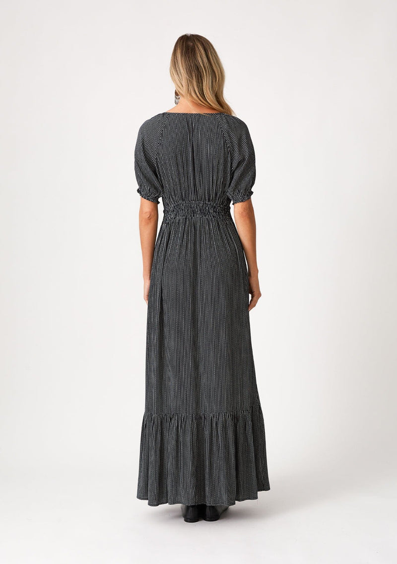 [Color: Navy] A back facing image of a blonde model wearing a best selling bohemian maxi dress in a navy blue dot print. With short puff sleeves, a v neckline, a long flowy tiered skirt with a side slit, and a half smocked elastic waist at the back. 