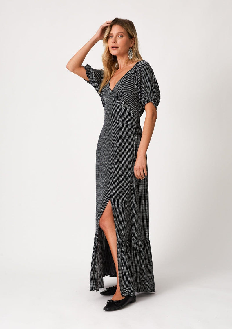 [Color: Navy] A full body side facing image of a blonde model wearing a best selling bohemian maxi dress in a navy blue dot print. With short puff sleeves, a v neckline, a long flowy tiered skirt with a side slit, and a half smocked elastic waist at the back. 