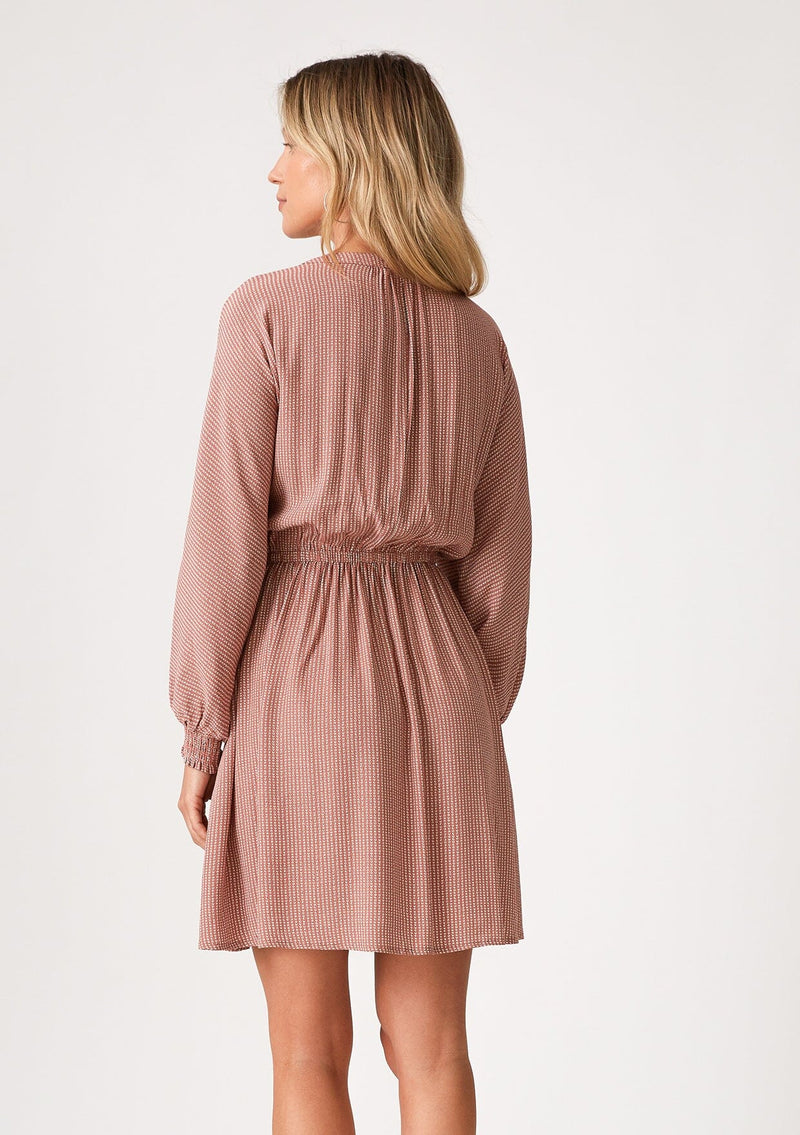 [Color: Mauve] A back facing image of a blonde model wearing a mauve pink dot print fall mini dress. With voluminous long sleeves, smocked elastic wrist cuffs, a v neckline, a self covered button front, and an elastic waist. 