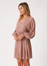 [Color: Mauve] A side facing image of a blonde model wearing a mauve pink dot print fall mini dress. With voluminous long sleeves, smocked elastic wrist cuffs, a v neckline, a self covered button front, and an elastic waist. 