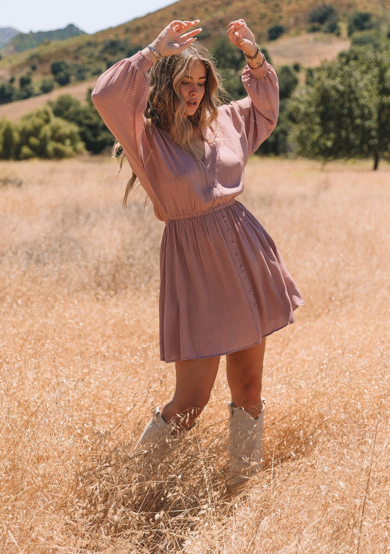 [Color: Mauve] A front facing image of a blonde model standing in a field wearing a mauve pink dot print fall mini dress. With voluminous long sleeves, smocked elastic wrist cuffs, a v neckline, a self covered button front, and an elastic waist.