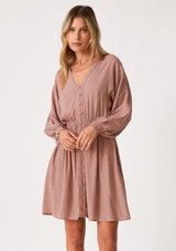 [Color: Mauve] A front facing image of a blonde model wearing a mauve pink dot print fall mini dress. With voluminous long sleeves, smocked elastic wrist cuffs, a v neckline, a self covered button front, and an elastic waist. 