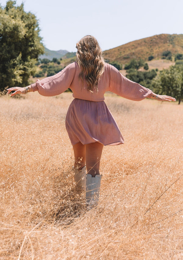 [Color: Mauve] A back facing image of a blonde model standing in a field wearing a mauve pink dot print fall mini dress. With voluminous long sleeves, smocked elastic wrist cuffs, a v neckline, a self covered button front, and an elastic waist.
