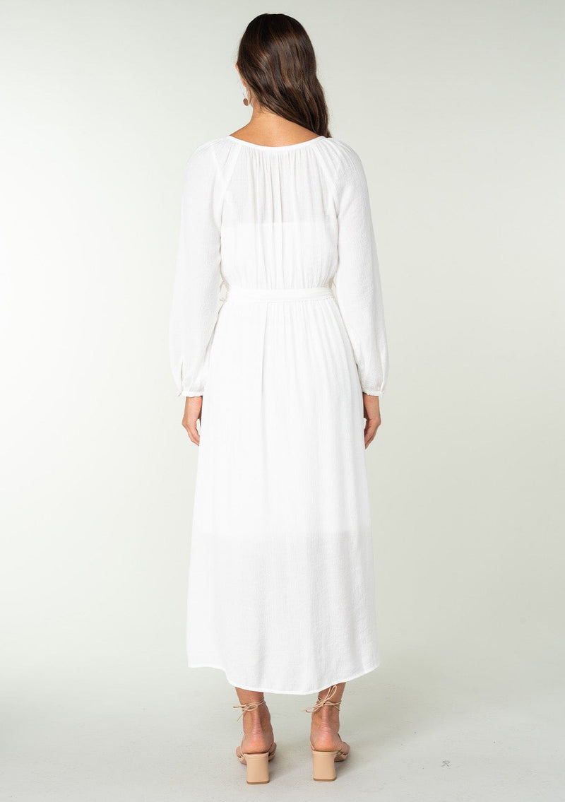 [Color: White] A back facing image of a brunette model wearing a classic white bohemian maxi dress. With long sleeves, a round neckline, a button front, side pockets, and a self tie waist belt.