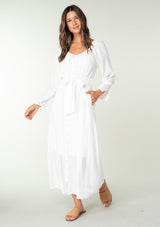 [Color: White] A front facing image of a brunette model wearing a classic white bohemian maxi dress. With long sleeves, a round neckline, a button front, side pockets, and a self tie waist belt.