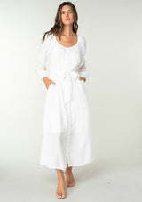 [Color: White] A front facing image of a brunette model wearing a classic white bohemian maxi dress. With long sleeves, a round neckline, a button front, side pockets, and a self tie waist belt.
