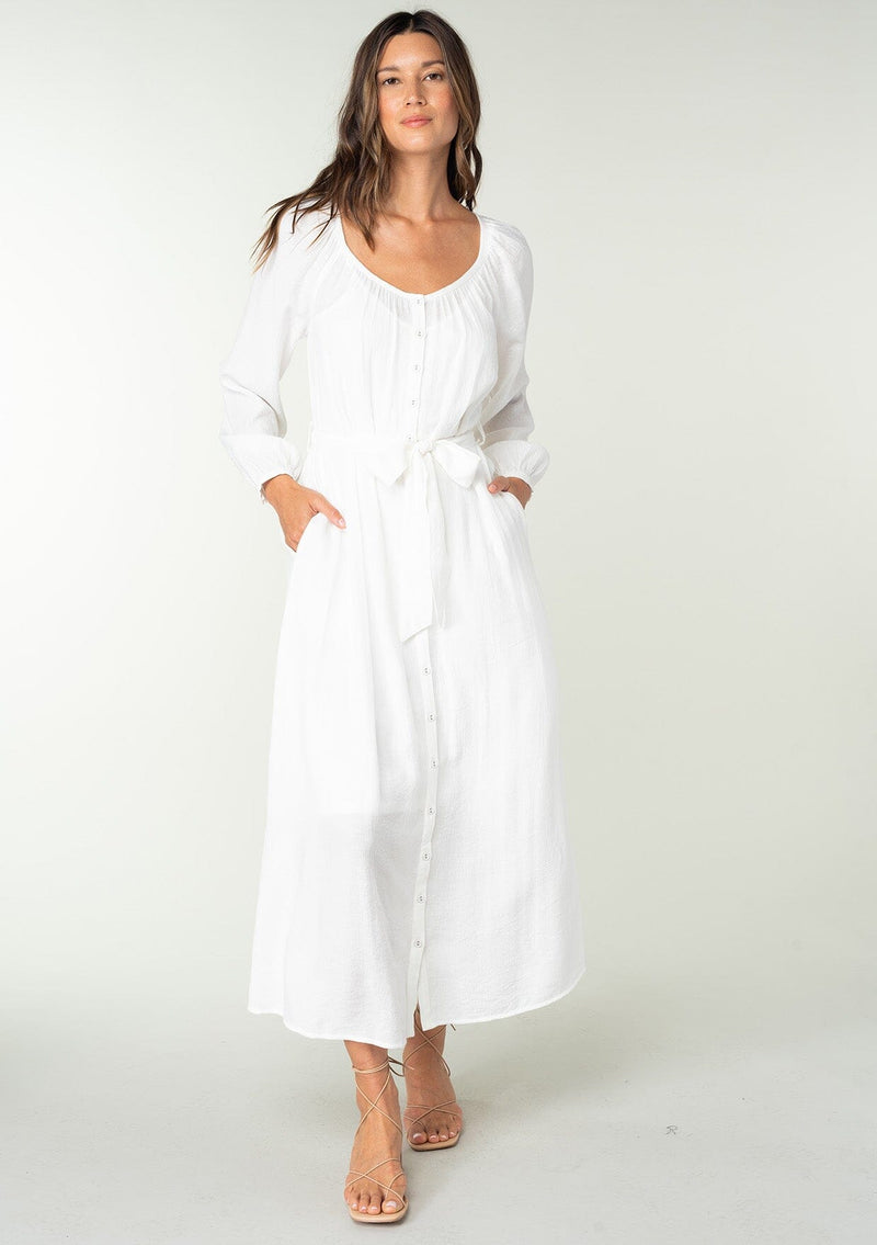 [Color: White] A front facing image of a brunette model wearing a classic white bohemian maxi dress. With long sleeves, a round neckline, a button front, side pockets, and a self tie waist belt.