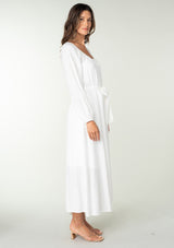 [Color: White] A side facing image of a brunette model wearing a classic white bohemian maxi dress. With long sleeves, a round neckline, a button front, side pockets, and a self tie waist belt.