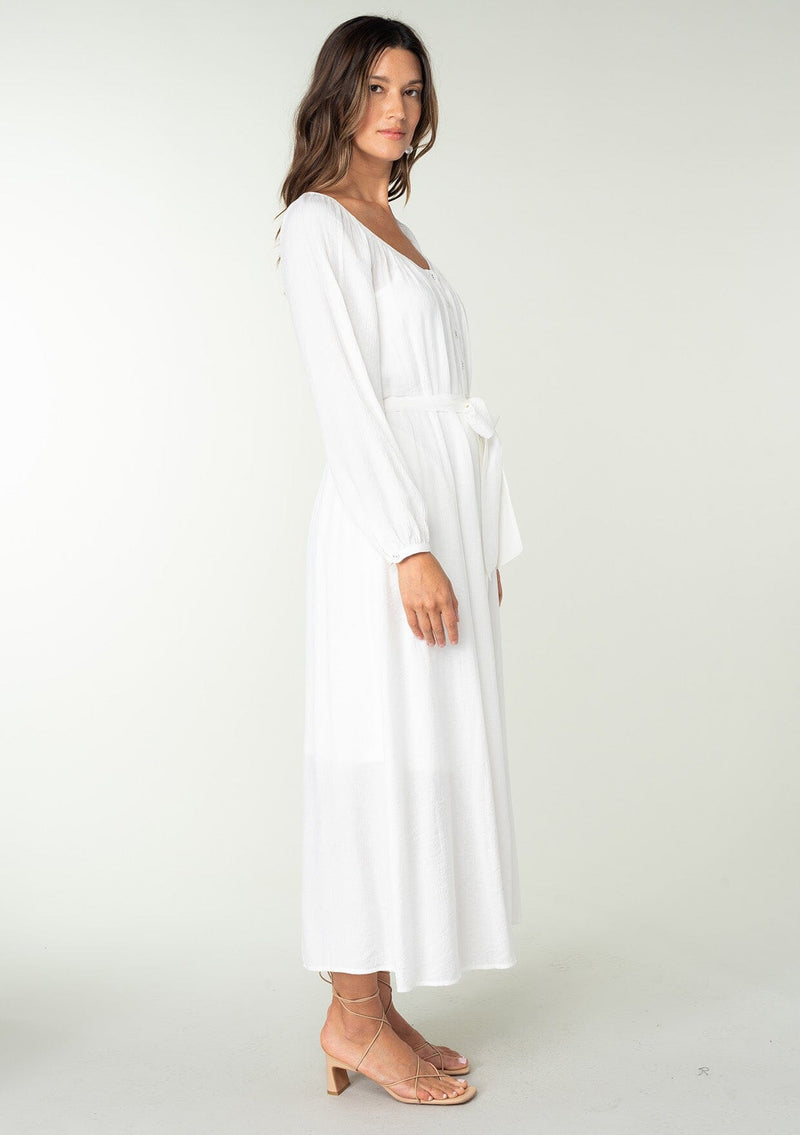 [Color: White] A side facing image of a brunette model wearing a classic white bohemian maxi dress. With long sleeves, a round neckline, a button front, side pockets, and a self tie waist belt.