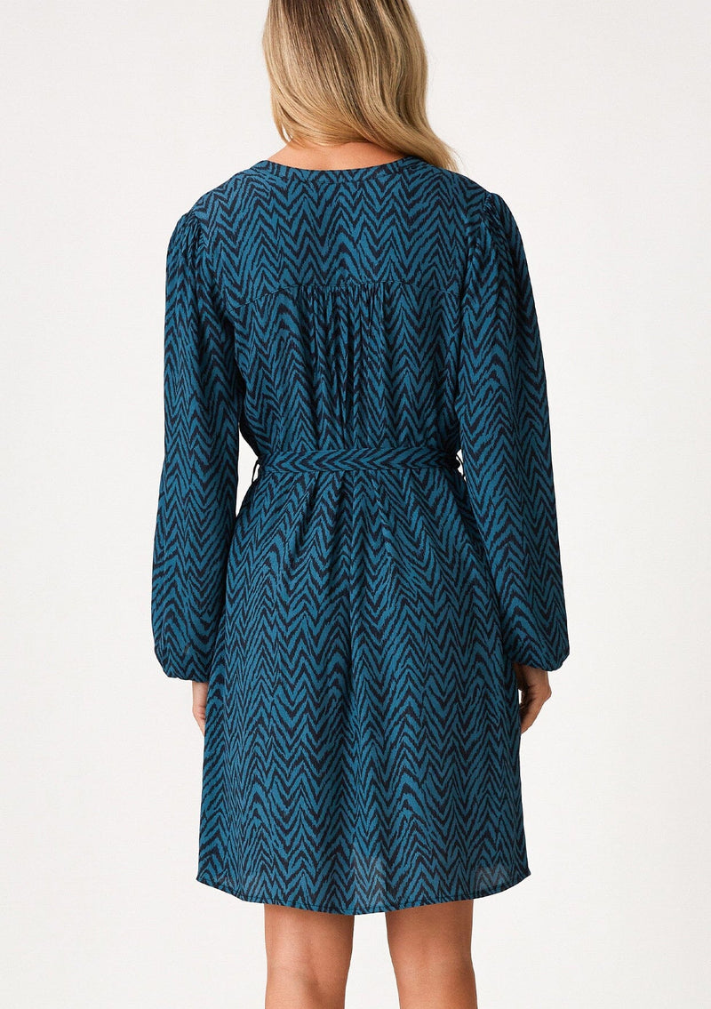 [Color: Teal/Navy] A back facing image of a blonde model wearing a bohemian fall mini dress in a teal and navy blue chevron stripe print. With voluminous long sleeves, a self covered button front, a v neckline, side pockets, and a self tie waist belt. 