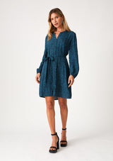[Color: Teal/Navy] A front facing image of a blonde model wearing a bohemian fall mini dress in a teal and navy blue chevron stripe print. With voluminous long sleeves, a self covered button front, a v neckline, side pockets, and a self tie waist belt. 
