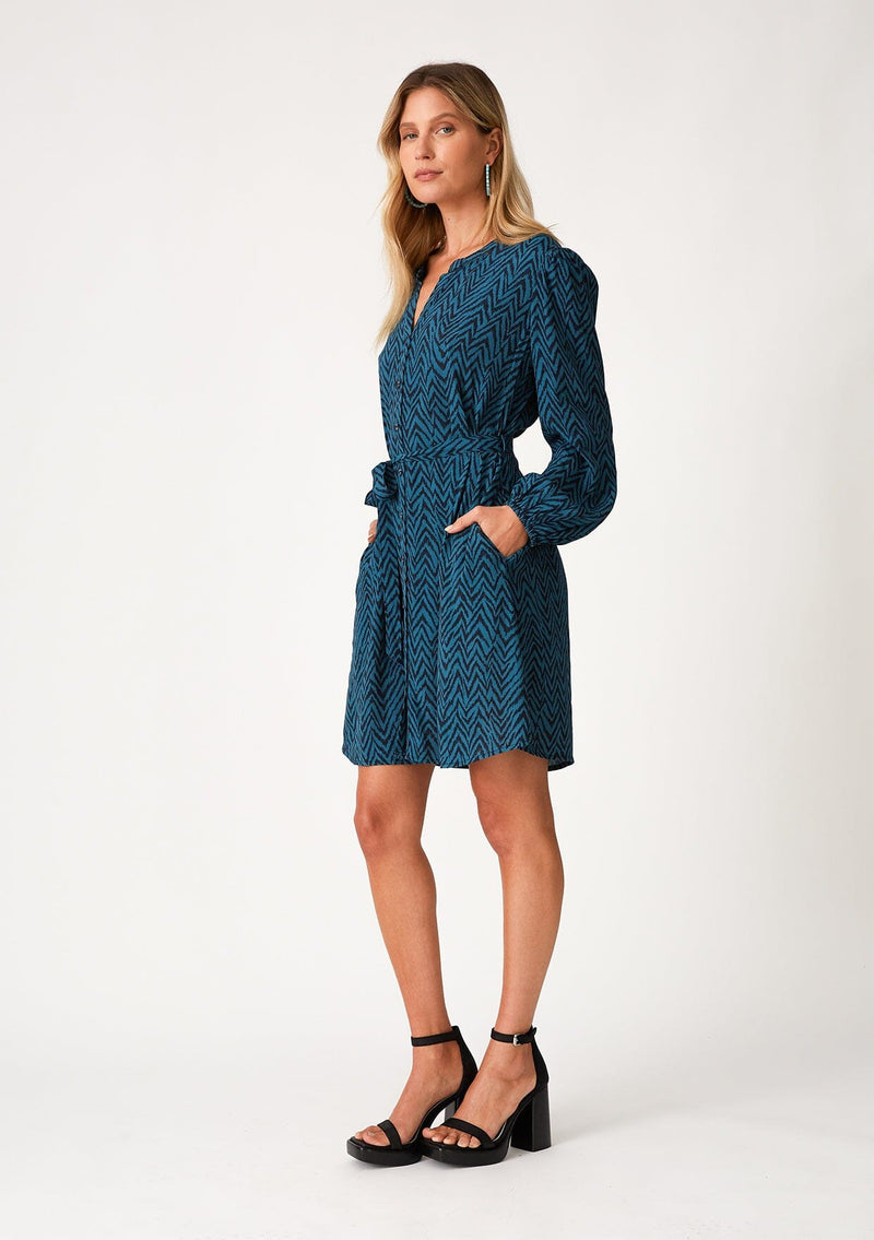 [Color: Teal/Navy] A full body side facing image of a blonde model wearing a bohemian fall mini dress in a teal and navy blue chevron stripe print. With voluminous long sleeves, a self covered button front, a v neckline, side pockets, and a self tie waist belt. 