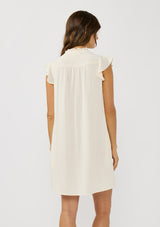 [Color: Tea] A back facing image of a brunette model wearing an off white button front mini dress with short flutter sleeves and side pockets. A relaxed and casual mini dress for the spring season 
