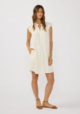 [Color: Tea] A front facing image of a brunette model wearing an off white button front mini dress with short flutter sleeves and side pockets. A relaxed and casual mini dress for the spring season 