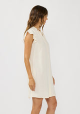 [Color: Tea] A side facing image of a brunette model wearing an off white button front mini dress with short flutter sleeves and side pockets. A relaxed and casual mini dress for the spring season 