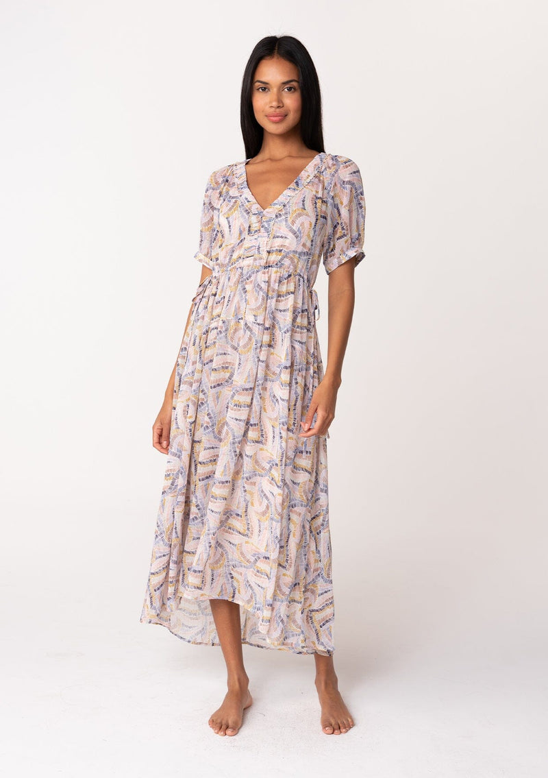 [Color: Natural/Mustard] A front facing image of a brunette model wearing a dreamy sheer chiffon maxi dress in a multi colored brush stroke print. With gold metallic thread details, short sleeves, a v neckline, a high low hemline, and an adjustable drawstring waist with side tie details. 
