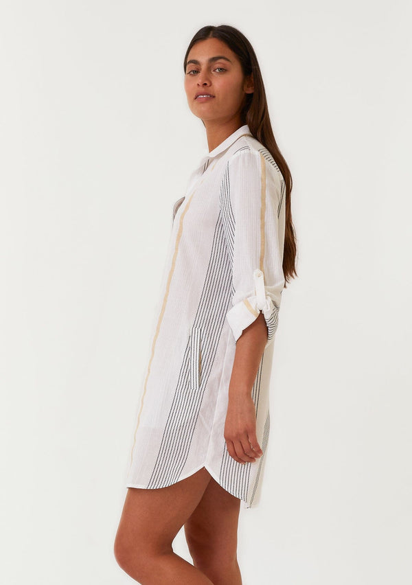 [Color: White/Gold] A side body front facing image of a brunette model wearing a white, black and gold striped mini shirt dress. A cotton spring dress with textured embroidered stripe details, a gold metallic lurex stripe, long rolled sleeves with a button tab closure, a split v neckline, a collared neckline, and side pockets. 