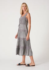 [Color: Grey] A side facing image of a blonde model wearing a grey bohemian mid length dress. With a square neckline, a tiered flowy skirt, adjustable tank top straps, a button front top, a drawstring tie waist, and lace trim throughout.