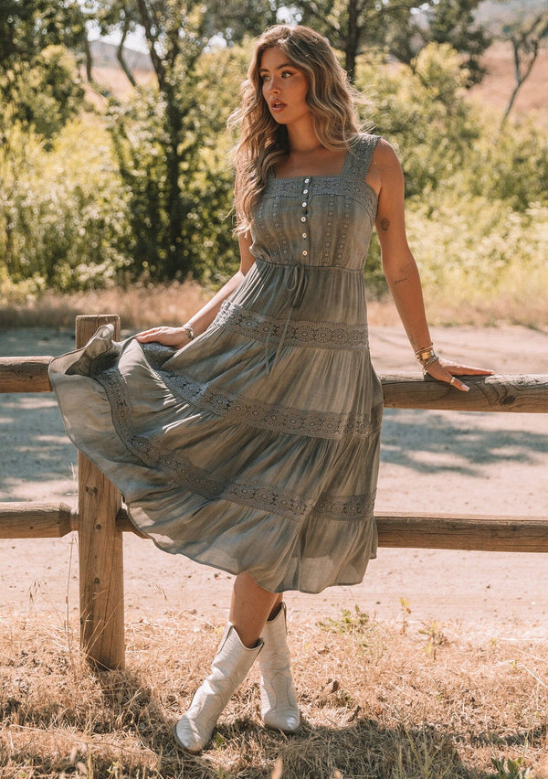 [Color: Grey] A full body front facing image of a blonde model standing outside wearing a grey bohemian mid length dress. With a square neckline, a tiered flowy skirt, adjustable tank top straps, a button front top, a drawstring tie waist, and lace trim throughout.