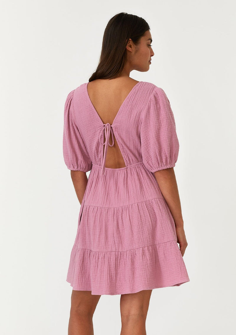 [Color: Orchid] A back facing image of a brunette model wearing a cute purple bohemian spring mini dress crafted in a soft cotton gauze. With short puff sleeves, a v neckline in the front and back, an empire waist, a tiered skirt, and an open back detail with a tie closure. 