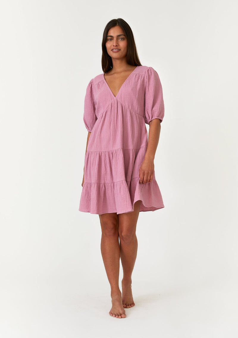 [Color: Orchid] A full body front facing image of a brunette model wearing a cute purple bohemian spring mini dress crafted in a soft cotton gauze. With short puff sleeves, a v neckline in the front and back, an empire waist, a tiered skirt, and an open back detail with a tie closure. 
