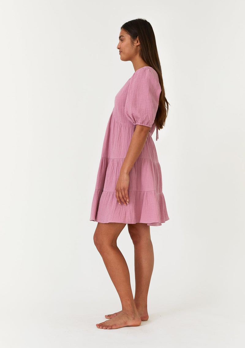 [Color: Orchid] A side facing image of a brunette model wearing a cute purple bohemian spring mini dress crafted in a soft cotton gauze. With short puff sleeves, a v neckline in the front and back, an empire waist, a tiered skirt, and an open back detail with a tie closure. 
