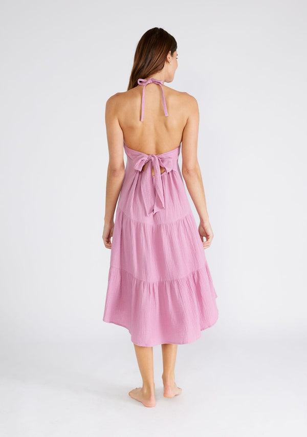 [Color: Orchid] A back facing image of a brunette model wearing a sleeveless halter mid length dress in a light purple cotton gauze. With an adjustable halter neckline, a tiered silhouette, a low back with adjustable tie closure, and a scooped neckline. 