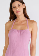 [Color: Orchid] A close up front facing image of a brunette model wearing a sleeveless halter mid length dress in a light purple cotton gauze. With an adjustable halter neckline, a tiered silhouette, a low back with adjustable tie closure, and a scooped neckline. 