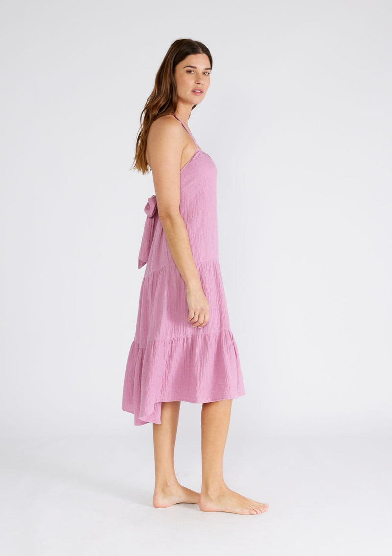 [Color: Orchid] A side facing image of a brunette model wearing a sleeveless halter mid length dress in a light purple cotton gauze. With an adjustable halter neckline, a tiered silhouette, a low back with adjustable tie closure, and a scooped neckline. 