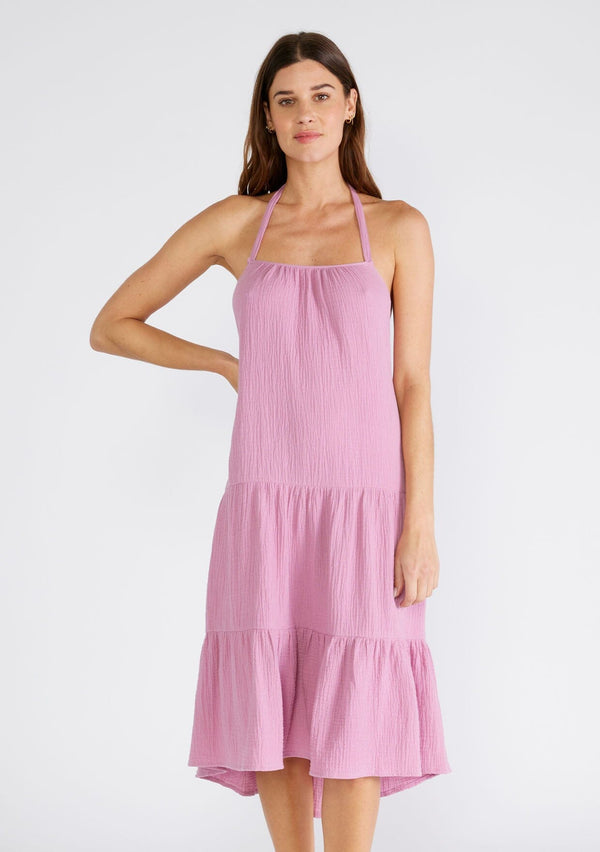 [Color: Orchid] A front facing image of a brunette model wearing a sleeveless halter mid length dress in a light purple cotton gauze. With an adjustable halter neckline, a tiered silhouette, a low back with adjustable tie closure, and a scooped neckline. 