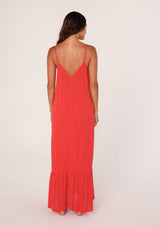 [Color: Flame] A back facing image of a brunette model wearing a simple flowy sleeveless maxi tank dress in a bright red crinkle rayon. With a v neckline in front and back, adjustable spaghetti straps, and a tiered skirt.