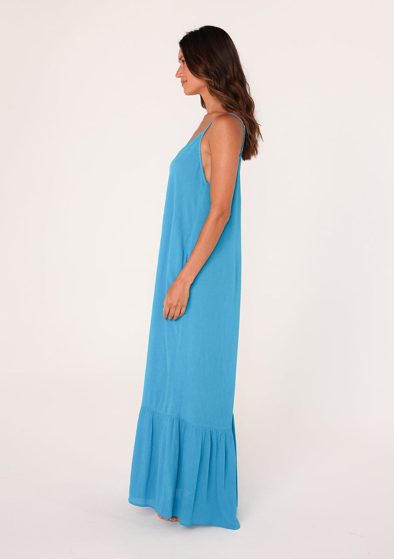 [Color: Turquoise] A side facing image of a brunette model wearing a simple flowy sleeveless maxi tank dress in a bright turquoise blue crinkle rayon. With a v neckline in front and back, adjustable spaghetti straps, and a tiered skirt.