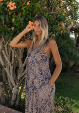[Color: Navy/Natural] An image of a blonde woman wearing a flowy bohemian floral print maxi dress standing outdoors with greenery and flowers in the background. 