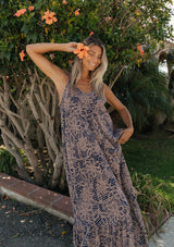 [Color: Navy/Natural] An image of a blonde woman wearing a flowy bohemian floral print maxi dress standing outdoors with greenery and flowers in the background. 