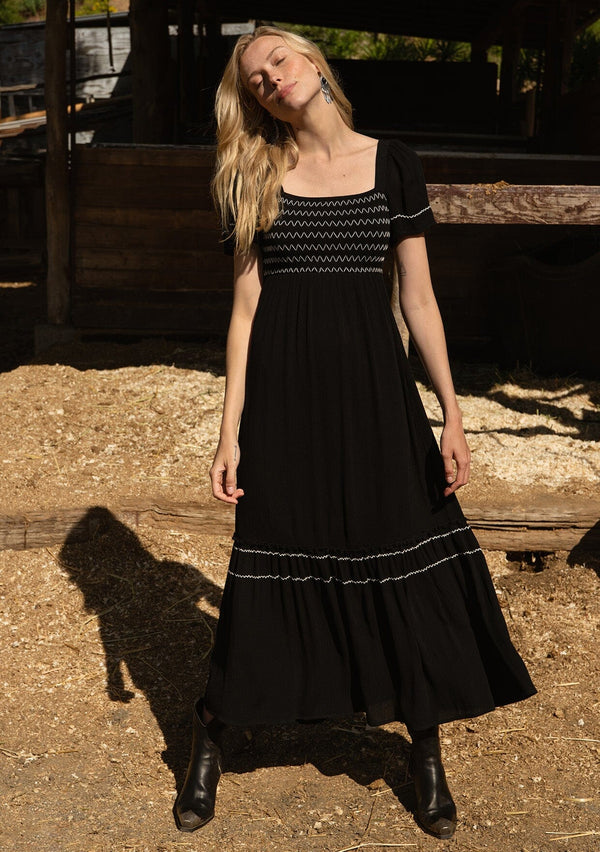 [Color: Black/Ivory] A front facing image of a blonde model standing outside at a ranch wearing a black bohemian maxi dress. With short sleeves, a slim fit smocked bodice, a square neckline in the front and back, a ruffle trimmed tiered skirt, side pockets, and contrast embroidered stitched detail.