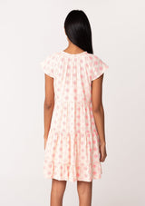 [Color: Natural/Pink] A back facing image of a brunette model wearing a summer cotton mini dress designed in pink embroidery. With short flutter cap sleeves, a split v neckline with tassel ties, a tiered silhouette, and a flowy relaxed fit. 