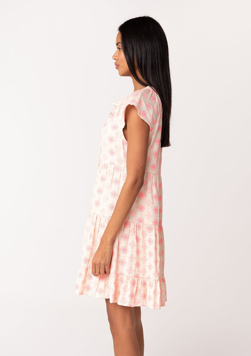 [Color: Natural/Pink] A side facing image of a brunette model wearing a summer cotton mini dress designed in pink embroidery. With short flutter cap sleeves, a split v neckline with tassel ties, a tiered silhouette, and a flowy relaxed fit. 