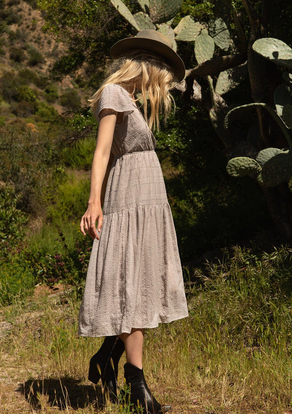[Color: Dove] A side facing image of a blonde model standing outside wearing a light grey bohemian spring mid length dress in all over eyelet lace chiffon. With short flutter sleeves, a surplice v neckline, a tiered flowy skirt with side slit, and an elastic waist.