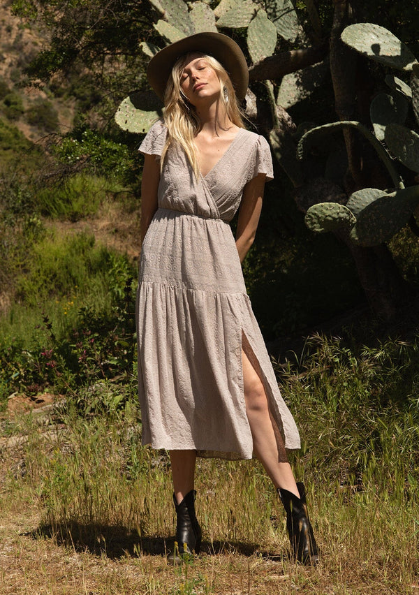 [Color: Dove] A front facing image of a blonde model standing outside wearing a light grey bohemian spring mid length dress in all over eyelet lace chiffon. With short flutter sleeves, a surplice v neckline, a tiered flowy skirt with side slit, and an elastic waist.