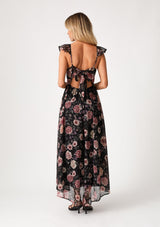 [Color: Black/Rose] A back facing image of a blonde model wearing a bohemian chiffon holiday maxi dress in a black and pink floral print. With a long flowy skirt, short flutter cap sleeves, adjustable spaghetti straps, a slim fit top, a scoop neckline, and an adjustable tie back detail with a sexy cutout. 