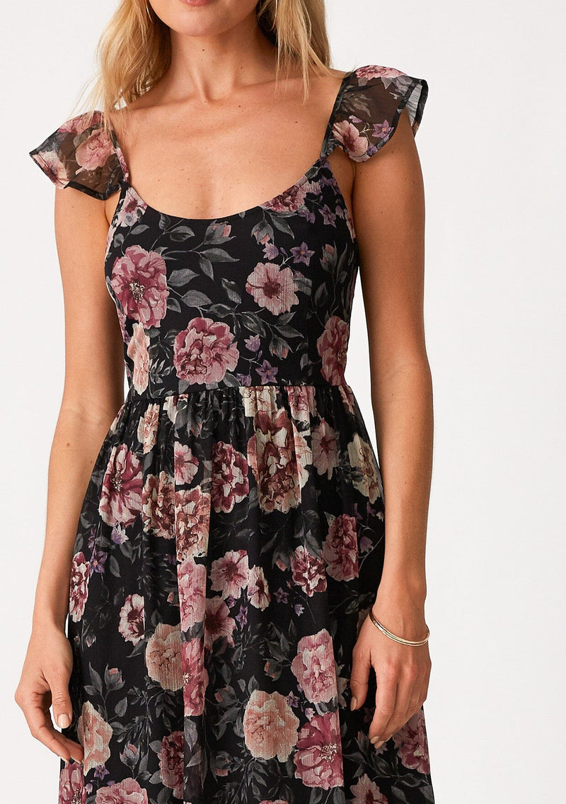 [Color: Black/Rose] A close up front facing image of a blonde model wearing a bohemian chiffon holiday maxi dress in a black and pink floral print. With a long flowy skirt, short flutter cap sleeves, adjustable spaghetti straps, a slim fit top, a scoop neckline, and an adjustable tie back detail with a sexy cutout. 