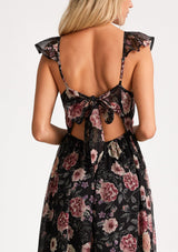 [Color: Black/Rose] A close up back facing image of a blonde model wearing a bohemian chiffon holiday maxi dress in a black and pink floral print. With a long flowy skirt, short flutter cap sleeves, adjustable spaghetti straps, a slim fit top, a scoop neckline, and an adjustable tie back detail with a sexy cutout. 