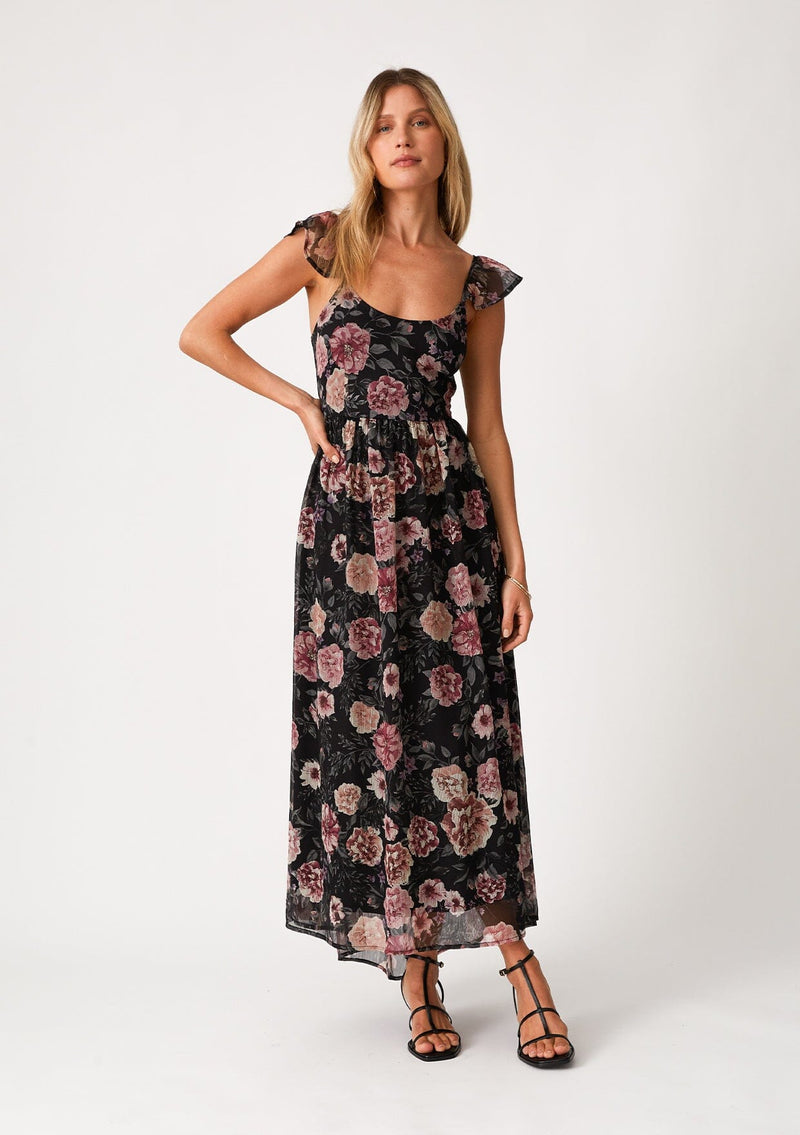 [Color: Black/Rose] A front facing image of a blonde model wearing a bohemian chiffon holiday maxi dress in a black and pink floral print. With a long flowy skirt, short flutter cap sleeves, adjustable spaghetti straps, a slim fit top, a scoop neckline, and an adjustable tie back detail with a sexy cutout. 