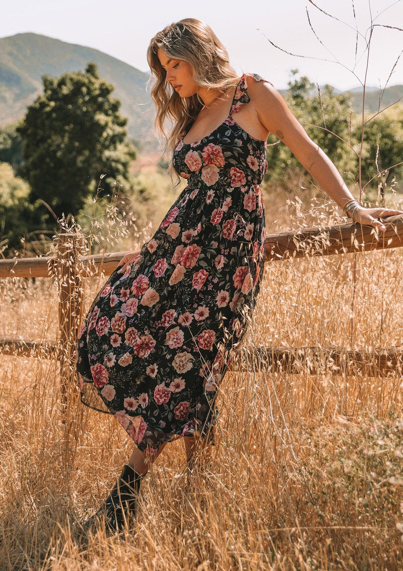[Color: Black/Rose] A side facing image of a blonde model standing in a field wearing a bohemian chiffon holiday maxi dress in a black and pink floral print. With a long flowy skirt, short flutter cap sleeves, adjustable spaghetti straps, a slim fit top, a scoop neckline, and an adjustable tie back detail with a sexy cutout.