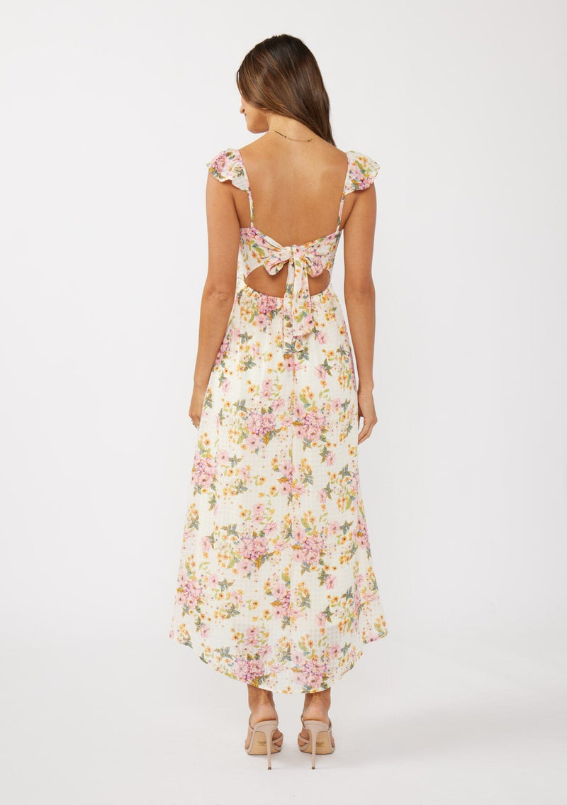 [Color: Natural/Dusty Pink] A back facing image of a brunette model in an off-white and pink floral maxi dress featuring flutter cap sleeves, a scoop neckline, an open back with tie detail, and a fit-and-flare silhouette. Ideal for Easter or special occasions.