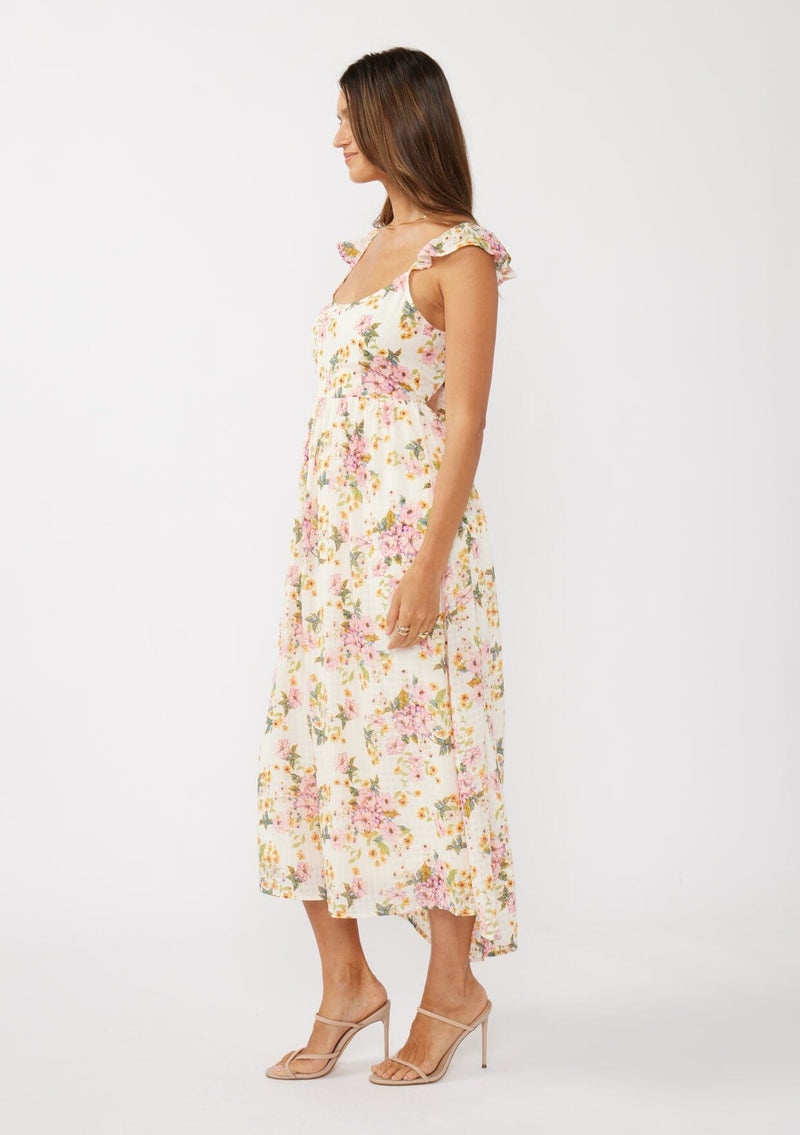 [Color: Natural/Dusty Pink] A side facing image of a brunette model in an off-white and pink floral maxi dress featuring flutter cap sleeves, a scoop neckline, an open back with tie detail, and a fit-and-flare silhouette. Ideal for Easter or special occasions.