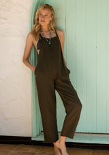 [Color: Military] A front facing image of a blonde model standing outside wearing a soft cotton gauze bohemian lounge jumpsuit in military green. With the silhouette of overalls, featuring tank top straps that attach with a button closure, two side pockets, a wide leg, and a racer back.