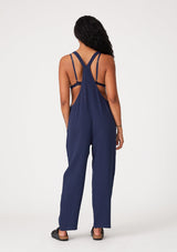 [Color: Royal] A back facing image of a brunette model wearing a soft cotton gauze bohemian lounge jumpsuit in royal blue. With the silhouette of overalls, featuring tank top straps that attach with a button closure, two side pockets, a wide leg, and a racer back.