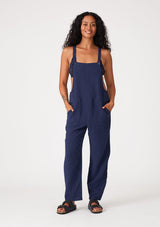 [Color: Royal] A full body front facing image of a brunette model wearing a soft cotton gauze bohemian lounge jumpsuit in royal blue. With the silhouette of overalls, featuring tank top straps that attach with a button closure, two side pockets, a wide leg, and a racer back.